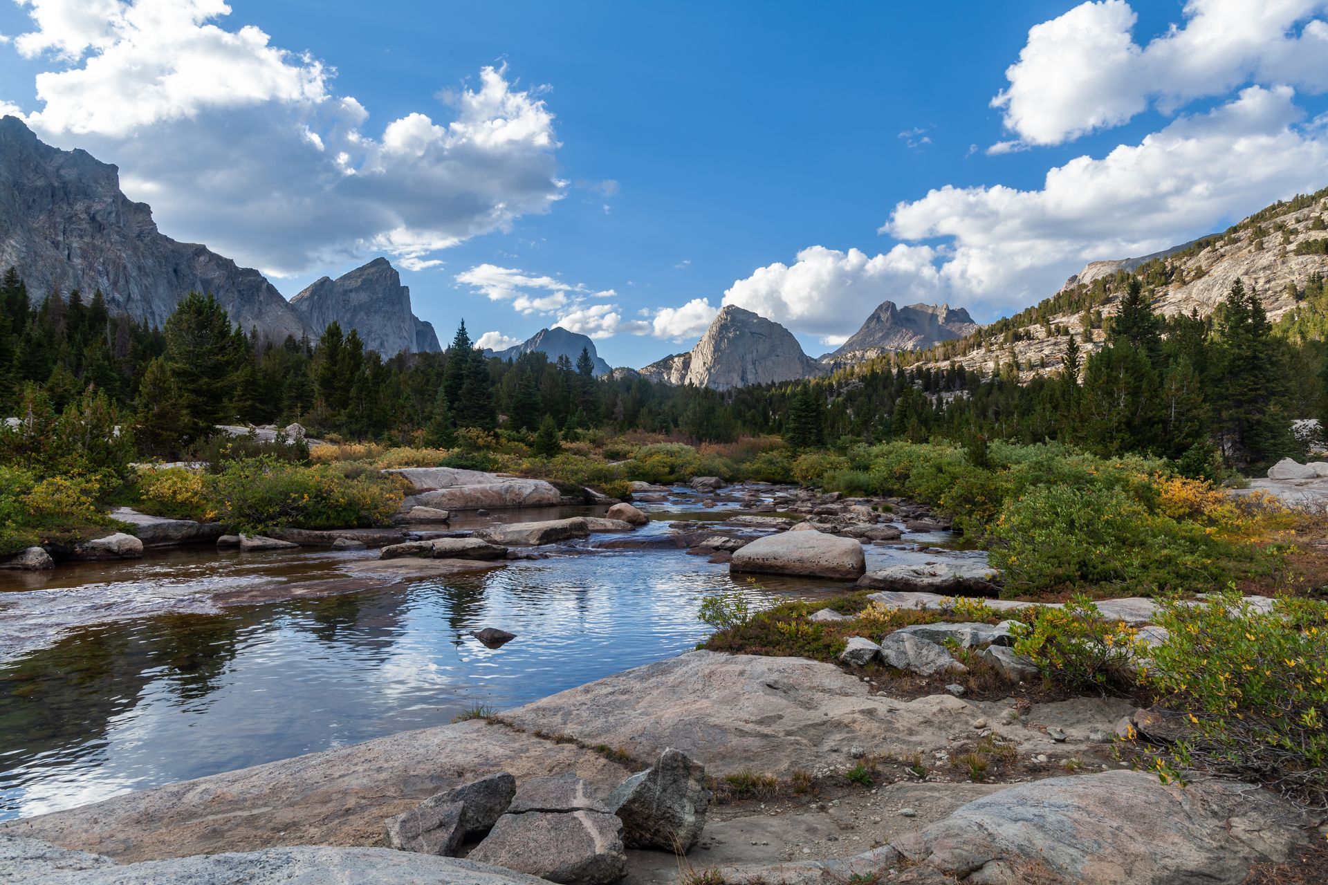 Clear stream in mountain valley, framed by rocky banks, green shrubs, and distant peaks under blue sky.