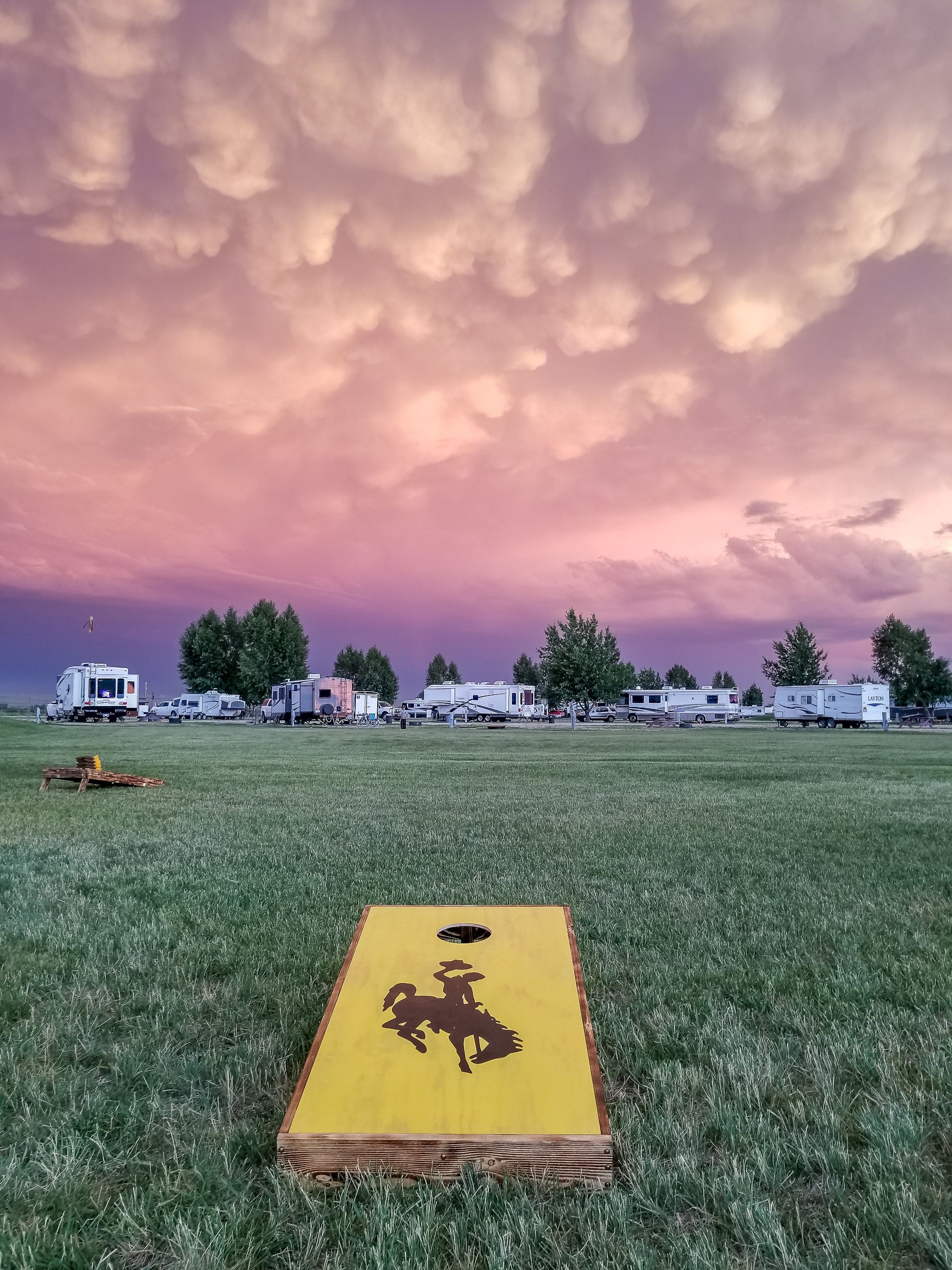 Cornhole board with cowboy graphic in a grassy field with trailers under a dramatic, colorful sunset.