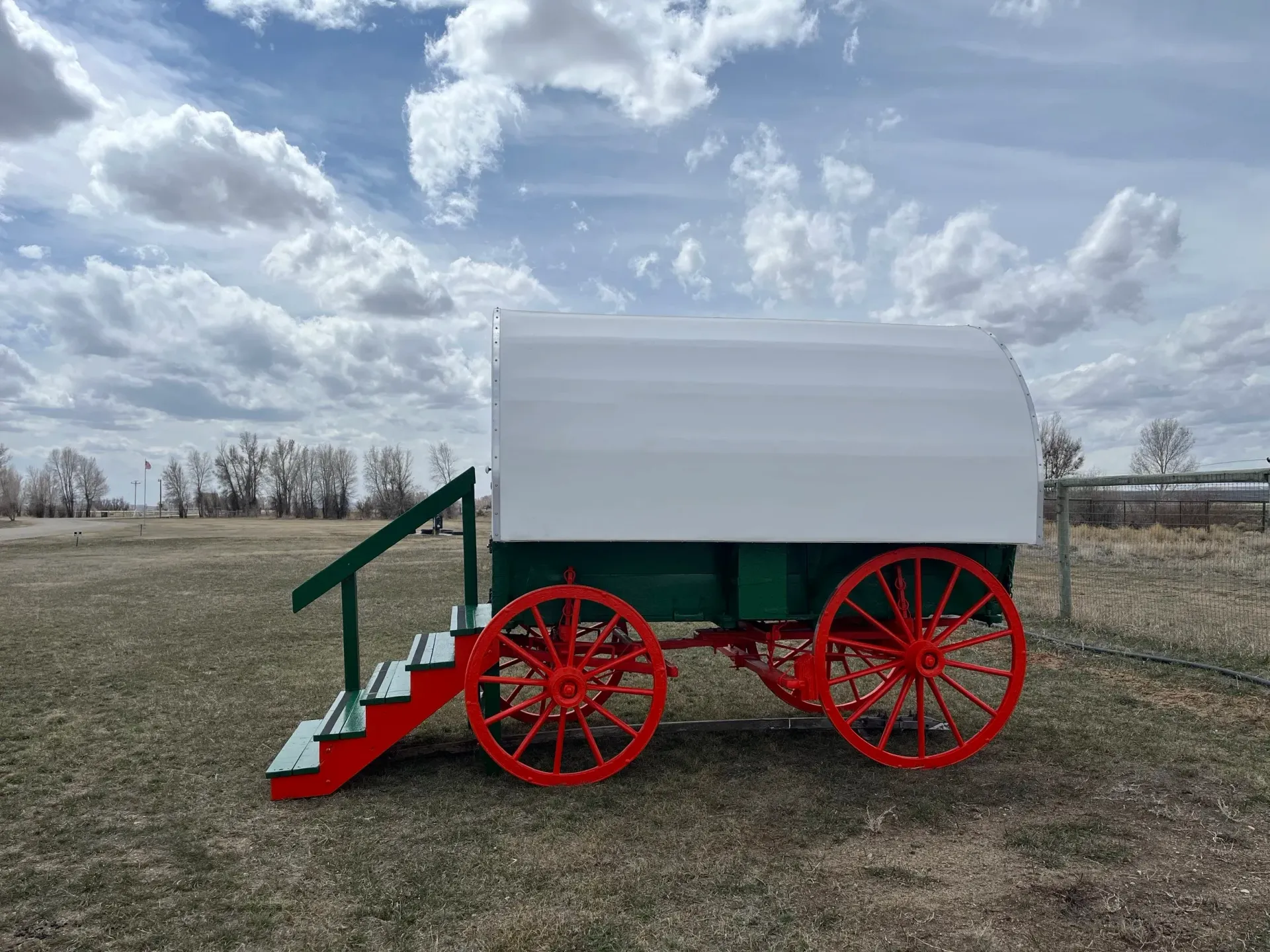 Red-wheeled wagon with a white top and green base sits on a grassy field under a cloudy sky.