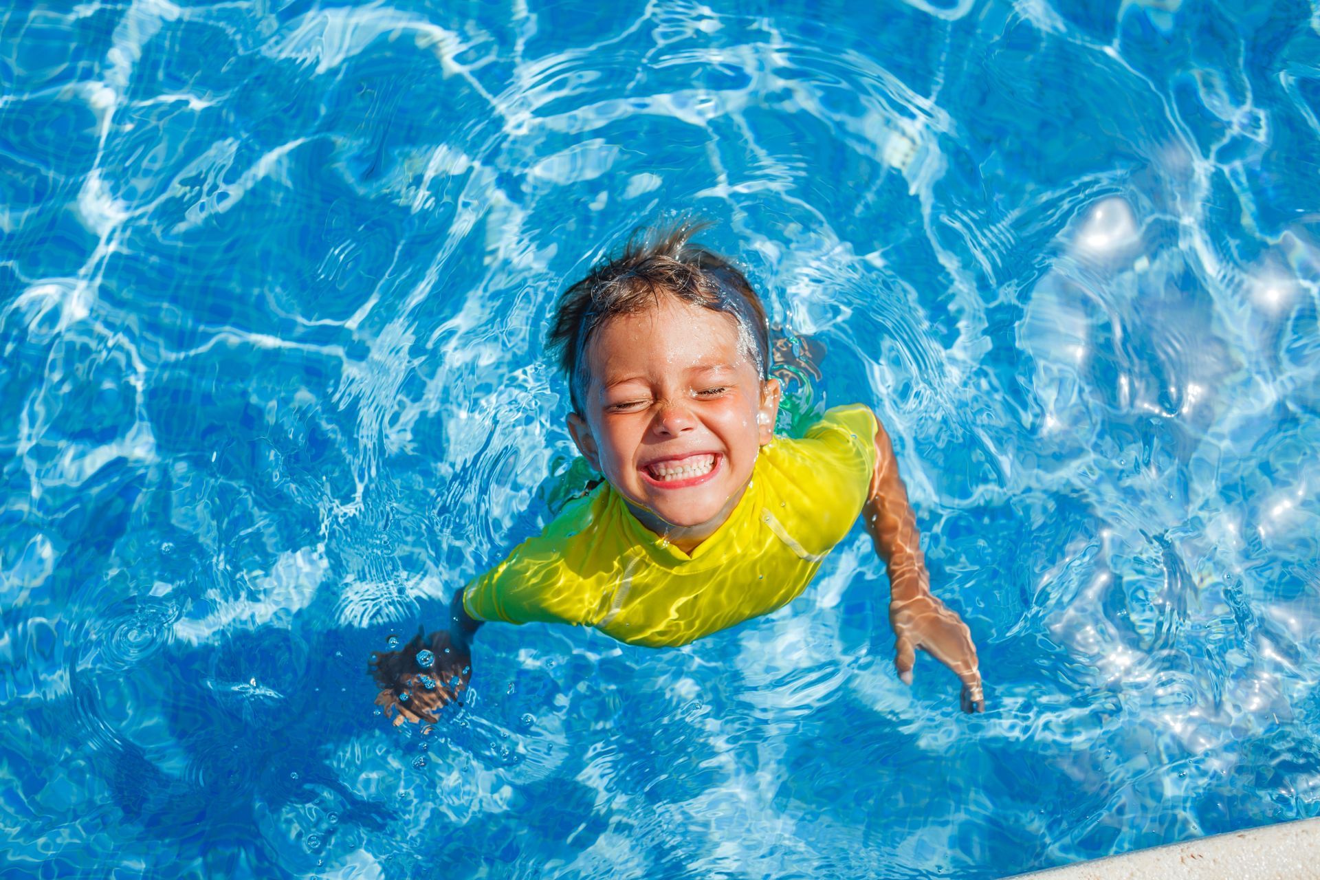 Smiling child floating in a bright blue swimming pool, wearing a yellow shirt