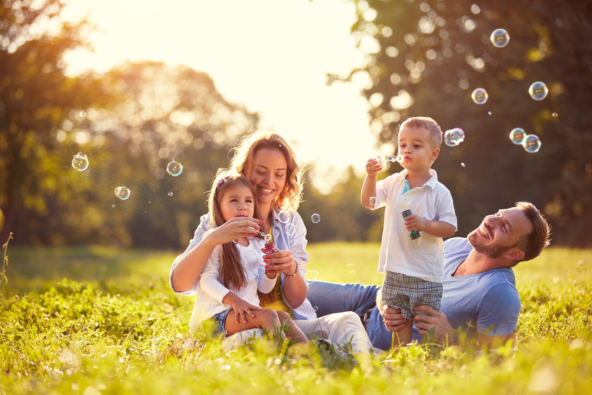 Four children sitting in sunlit grass, blowing bubbles and laughing in a park