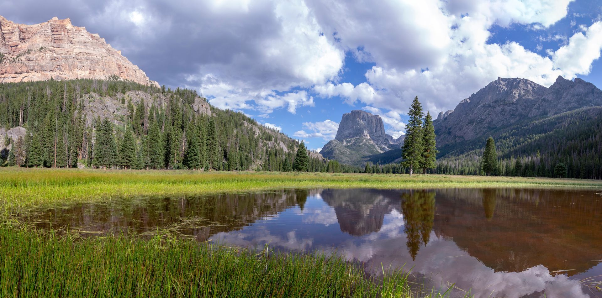 Panoramic view of a reflective pond in a grassy meadow surrounded by pine forests and dramatic rocky mountains.
