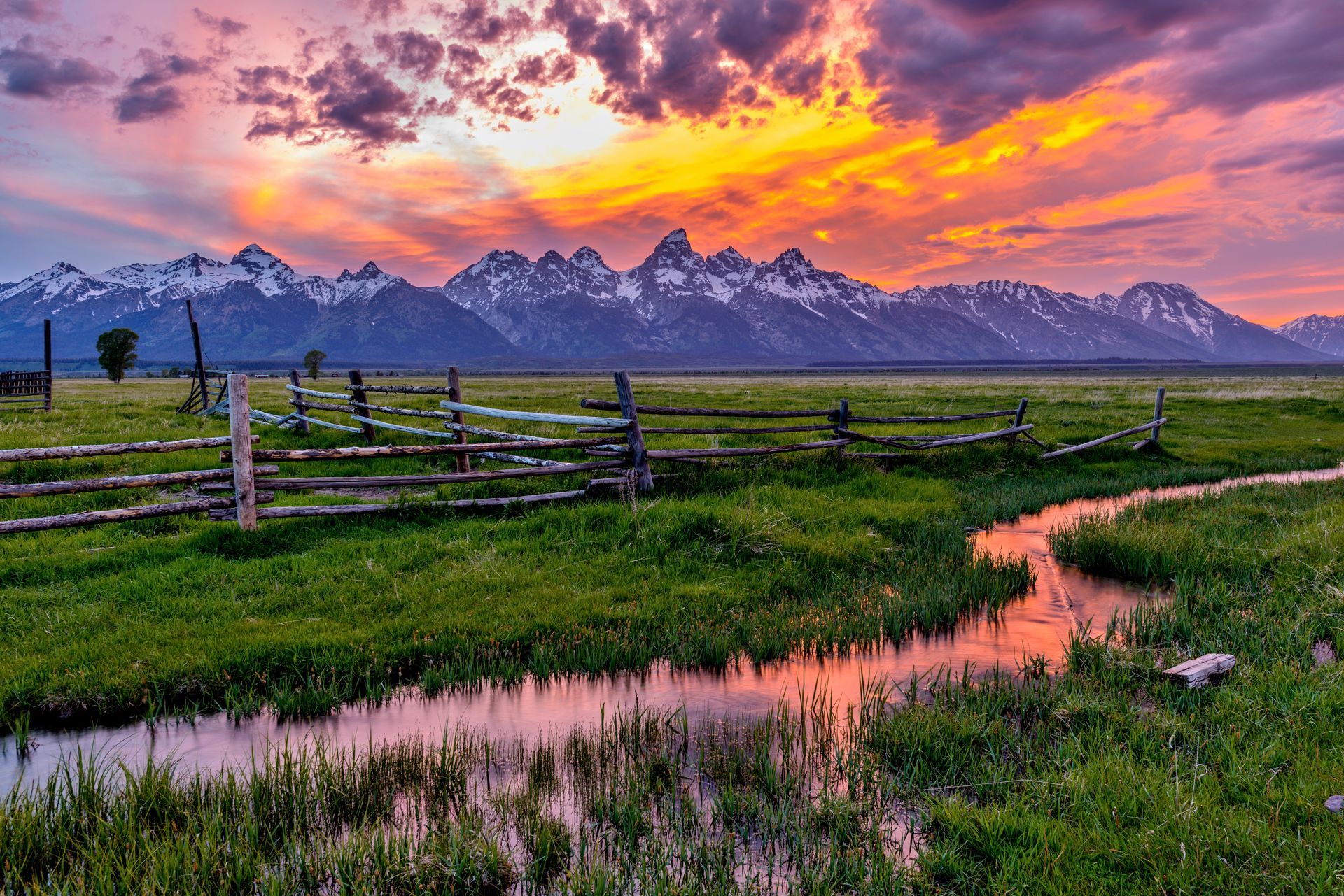 Sunset over snow-capped mountains and green fields, with a creek and wooden fence.