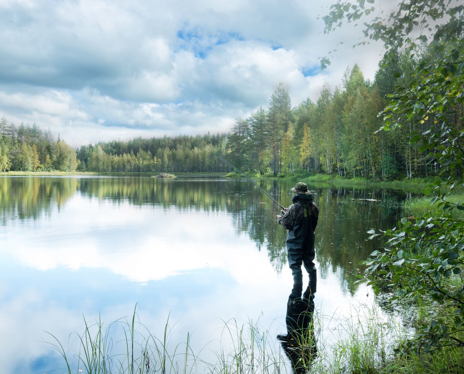 Man fishing in lake, reflection in water. Forest and cloudy sky in background.