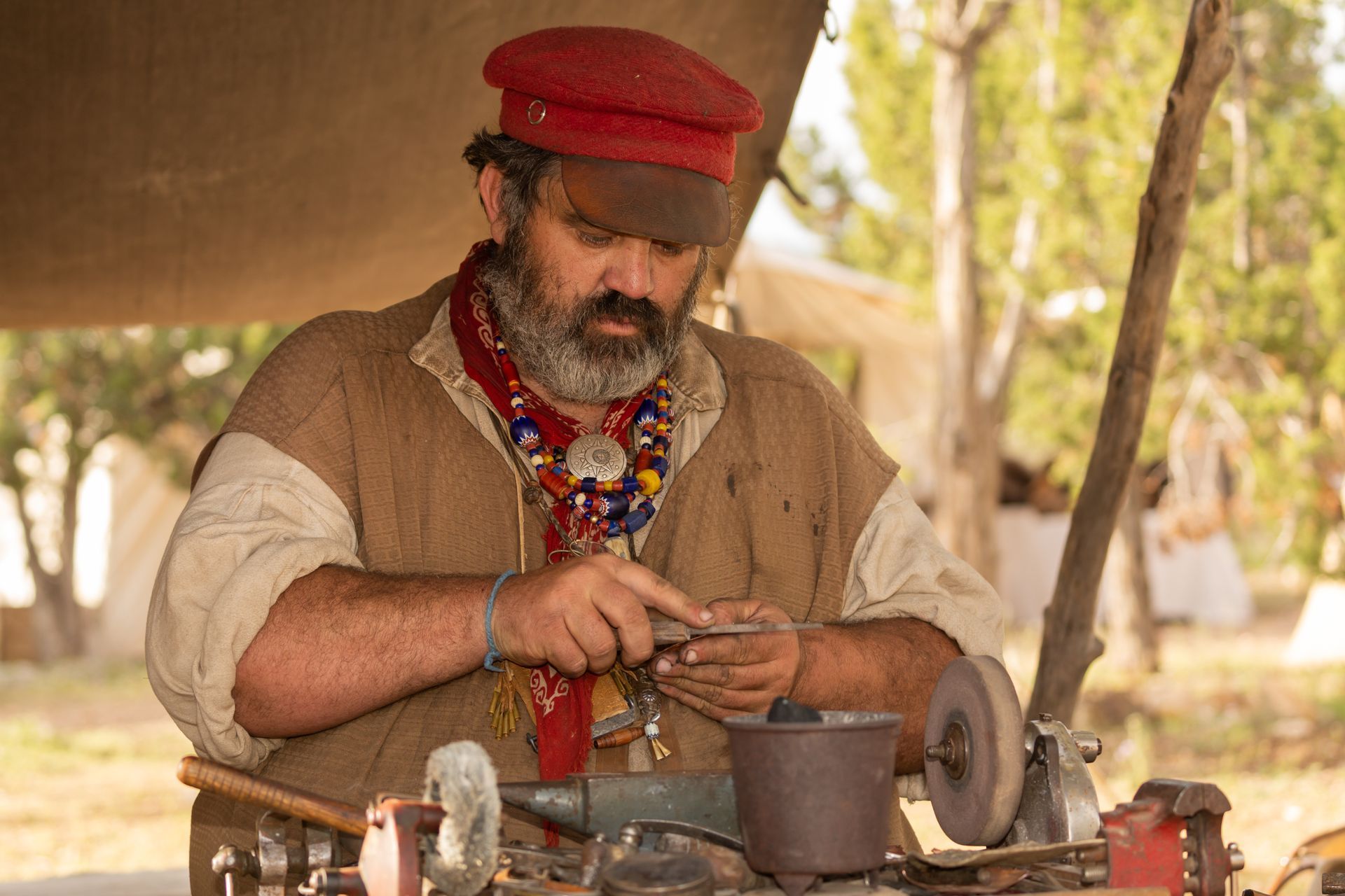 A person in a red cap and brown vest works at a small grinder, focusing intently on a tool in their hands.