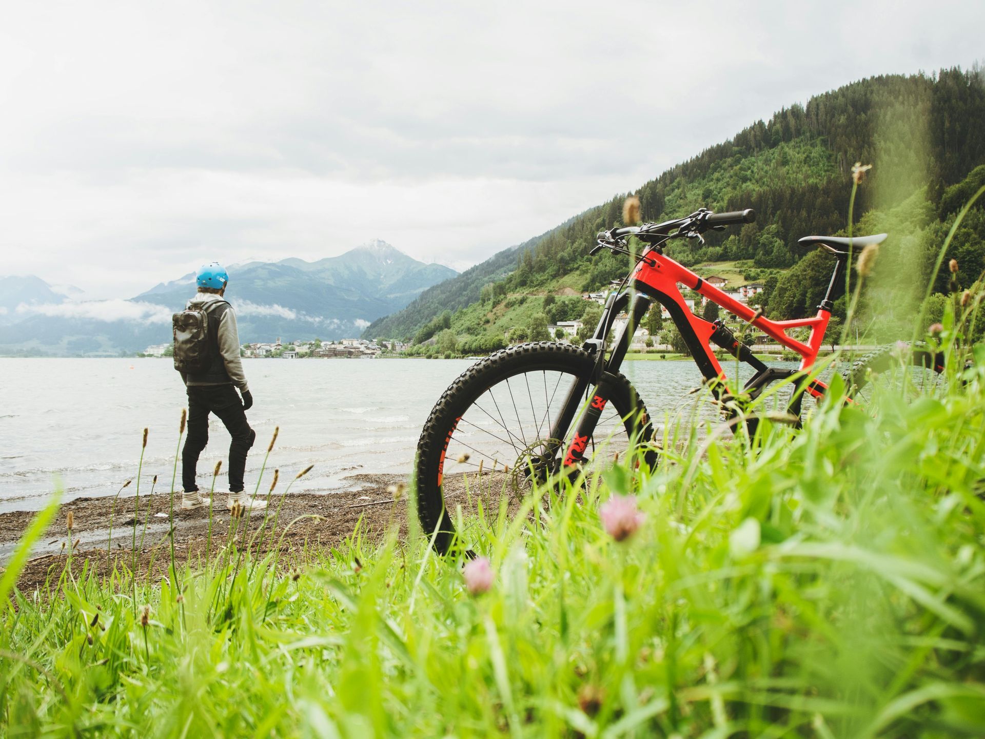 Person standing by lake with mountain bike, blue helmet, and backpack, overlooking a lake, mountains in background.