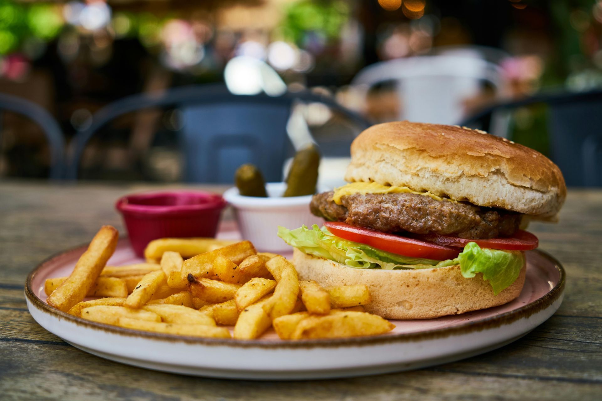 Burger with fries, pickles, and dipping sauce on a plate, outdoors on a wooden table.