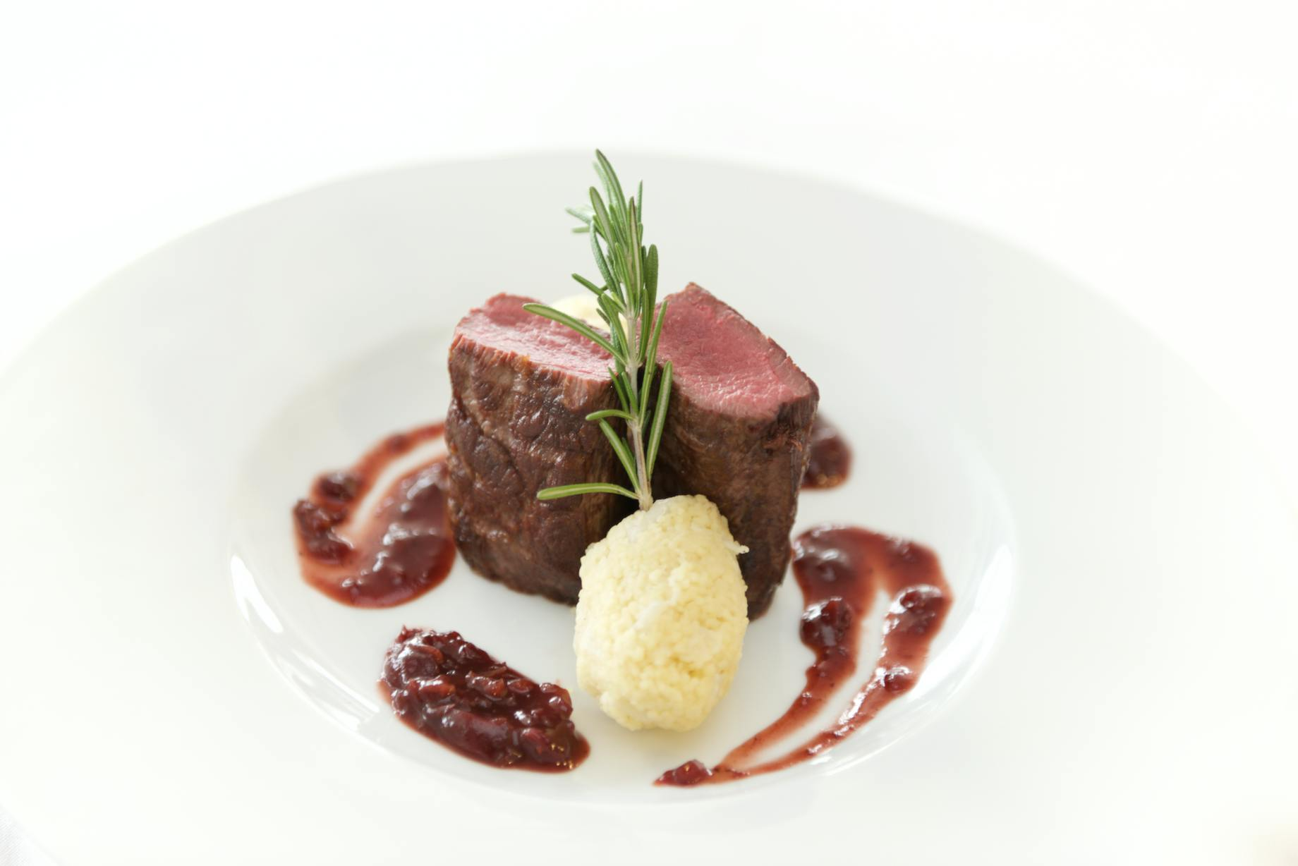 Steak medallions with red sauce and mashed potatoes, garnished with rosemary, on a white plate.