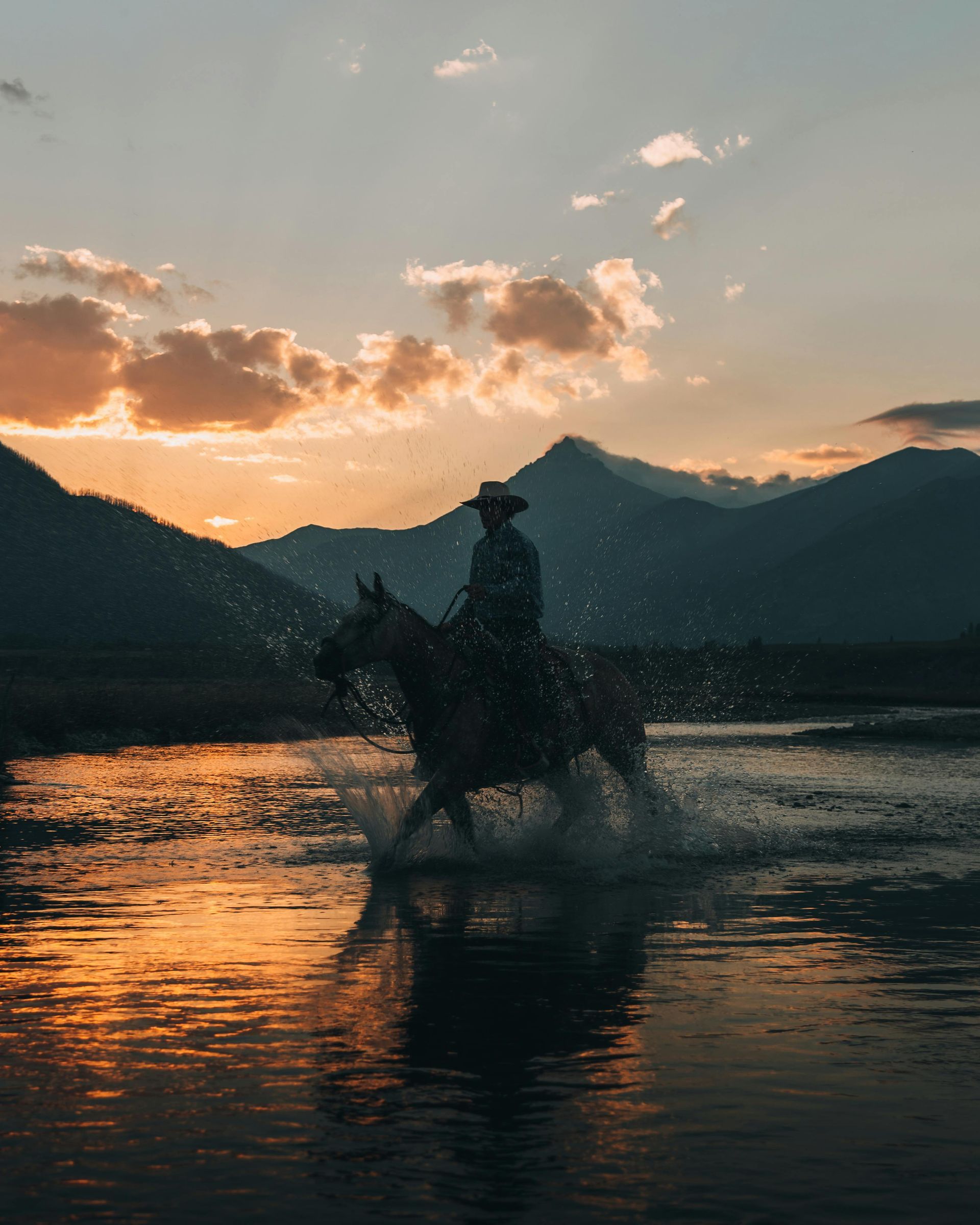 Cowboy on horseback riding through water at sunset, mountains in the background.