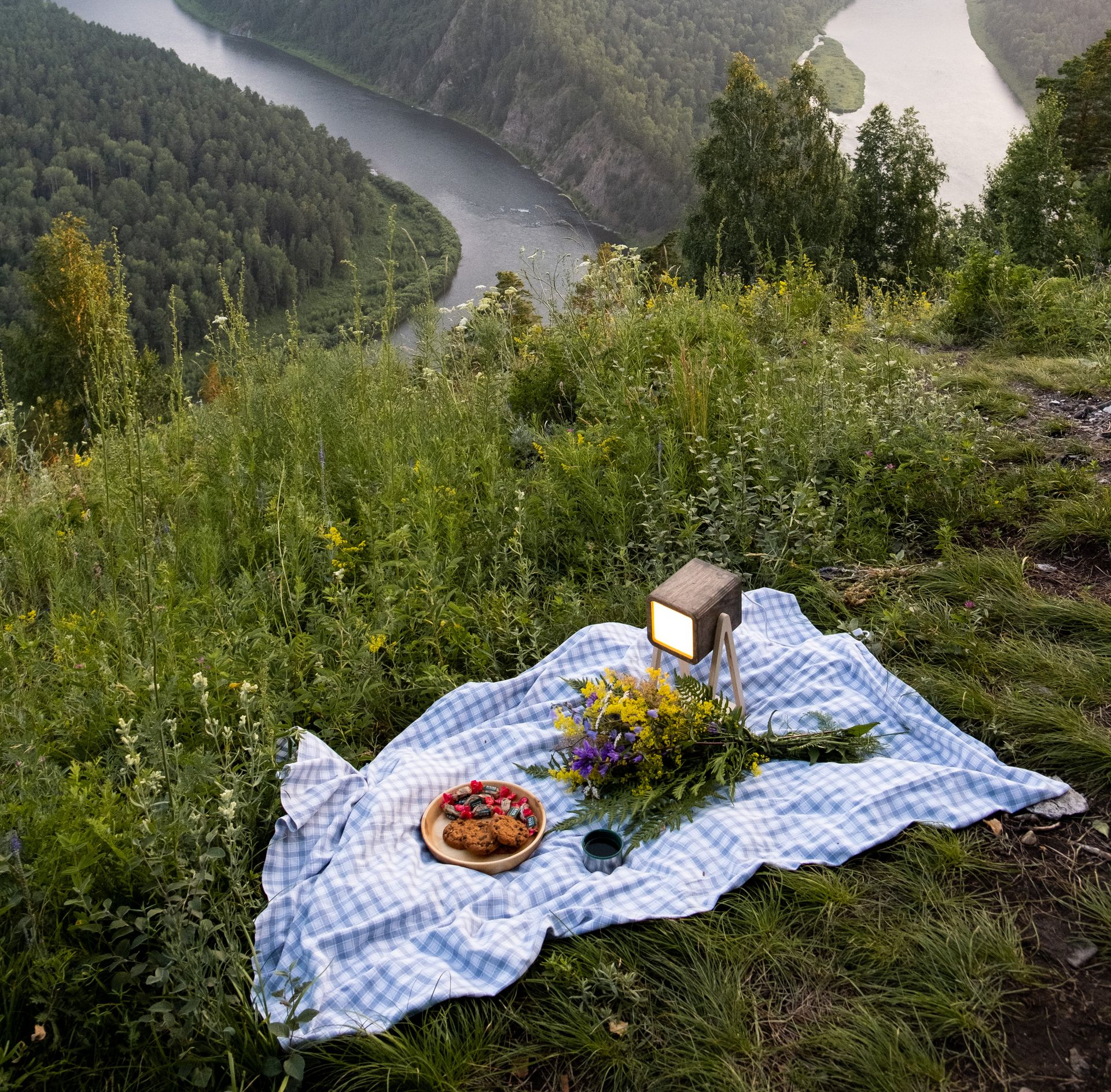 Picnic setup on a hillside overlooking a winding river. Blue checkered blanket, food, flowers, and a lantern.