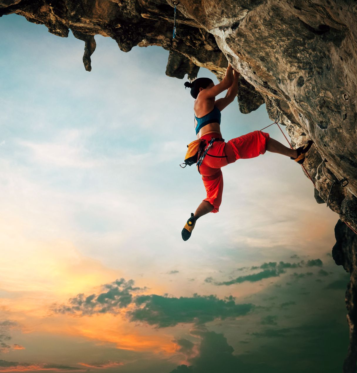 Rock climber ascending a steep cliff at sunset. Red pants, blue top. Sky with orange and blue hues.