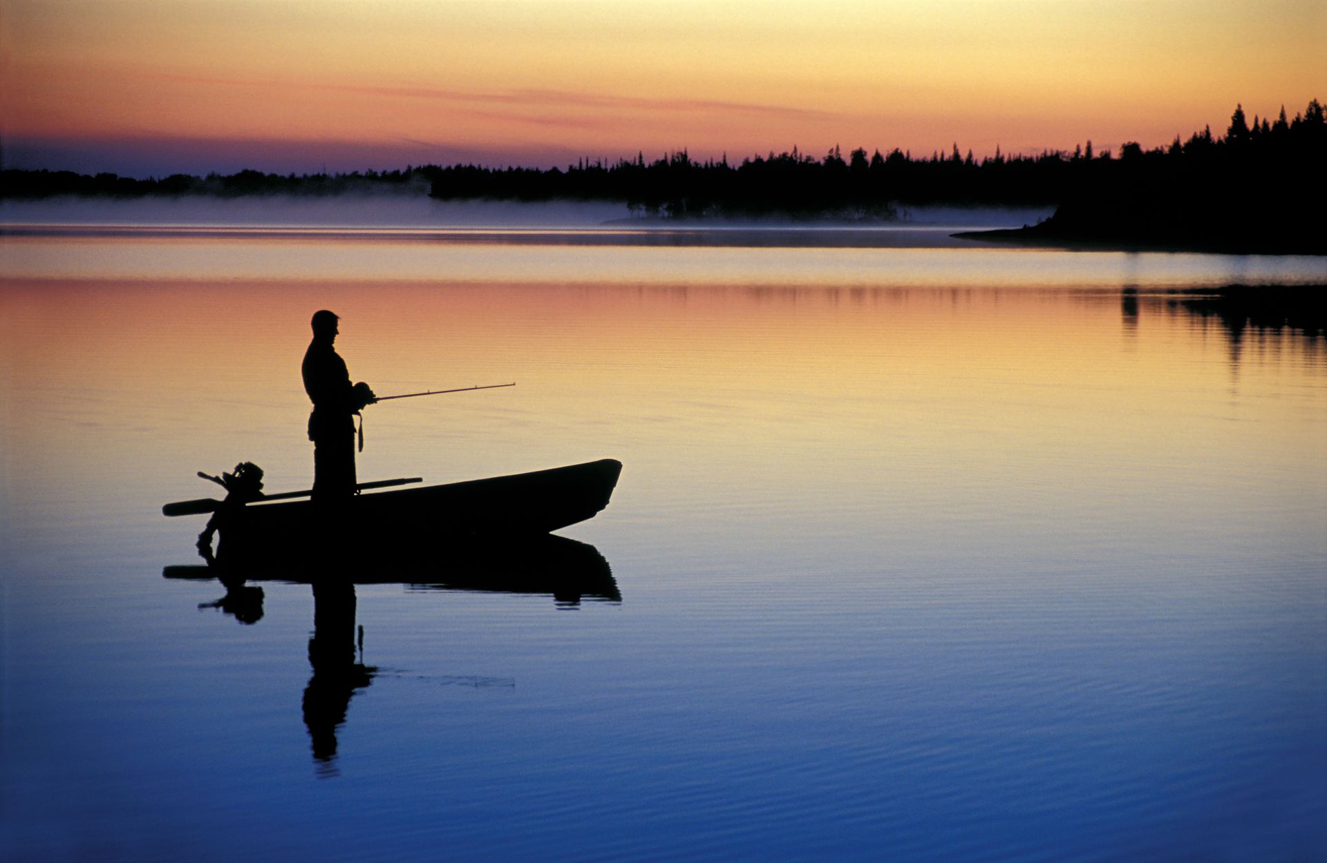 Silhouette of a person fishing from a boat on a calm lake at dusk; colorful sky reflected in water.