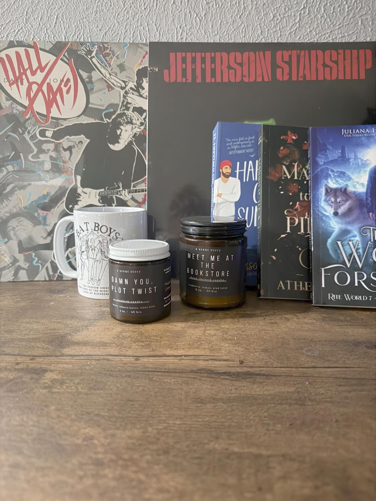 Two candles, mug, and books on a wooden surface in front of album art.
