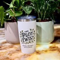 A white tumbler with "The Plot Twist" logo sits in front of potted plants.