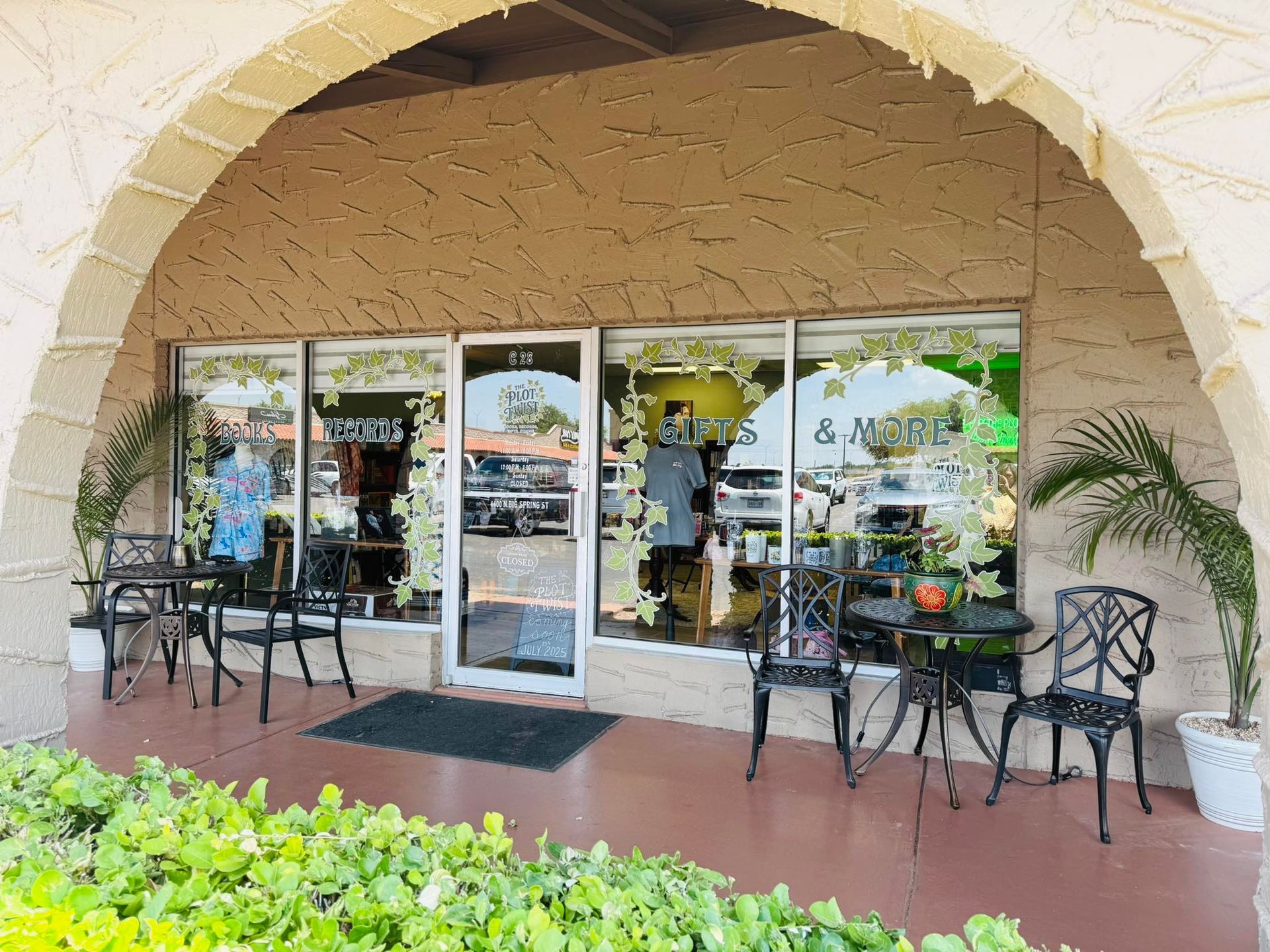 Storefront under an archway, with glass windows, tables, chairs, and potted plants.