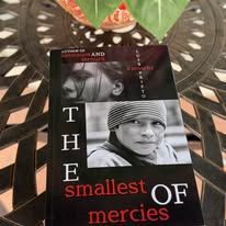 Book cover on a metal table, title "The Smallest of Mercies," faces of two people depicted.