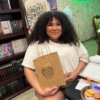 Woman holding a brown paper bag with text, smiling, in front of a bookshelf.