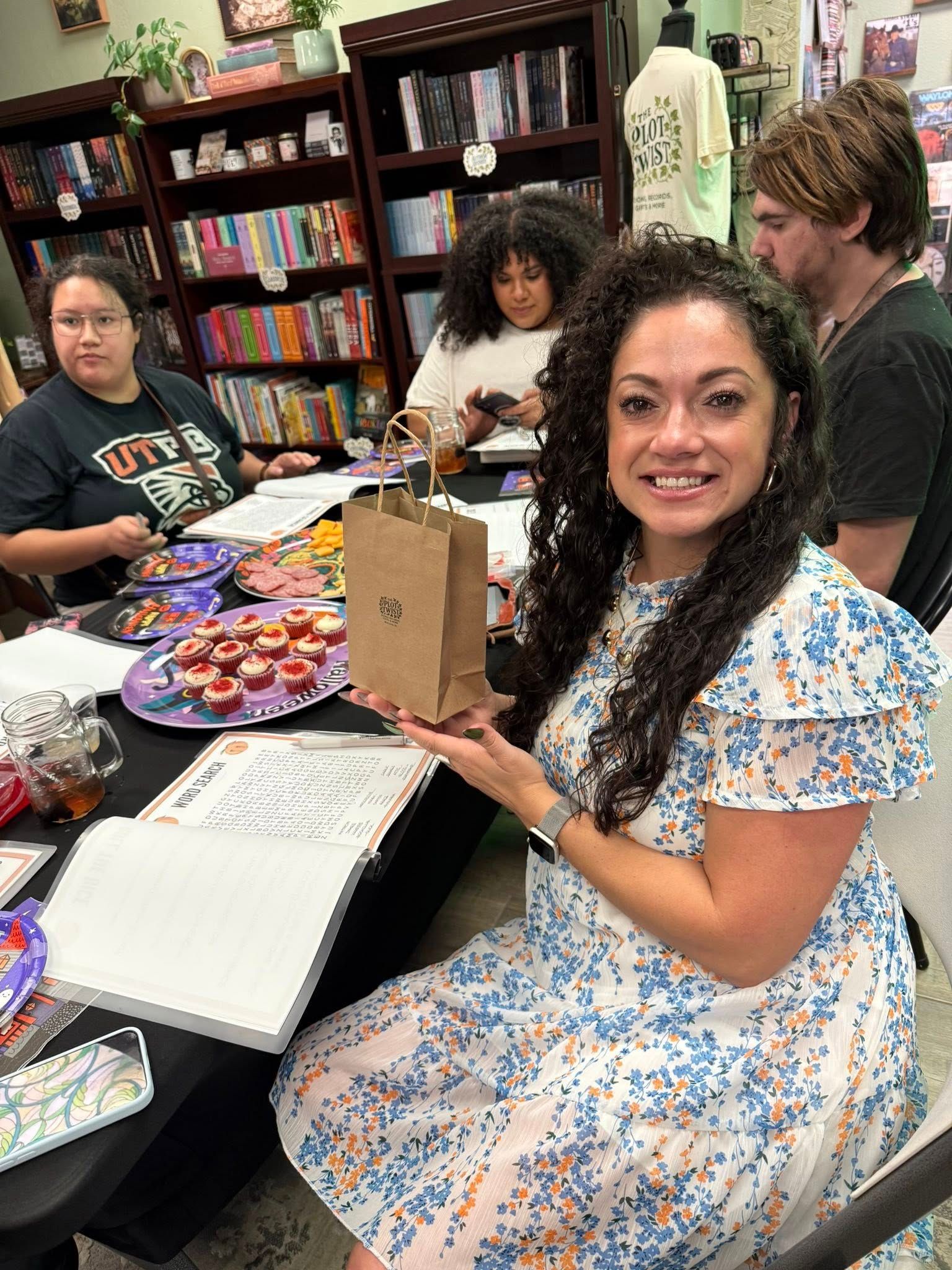 Woman holding small brown bag, smiling at a table with others. Snacks and books in the background.