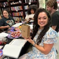 Woman smiles, holding a gift bag at a table with books and food. Others work/eat nearby.