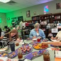 People at a table with food, drinks, and books. A woman is writing. Green and purple lighting.