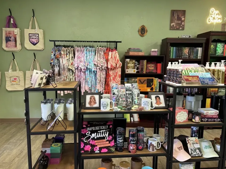 A retail store interior displays tote bags, clothing, books, mugs, and other merchandise on shelves and racks.