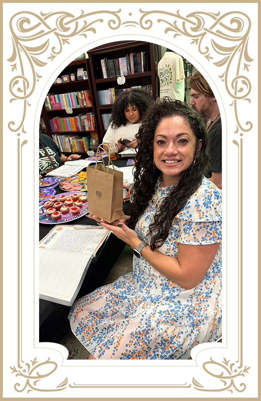 Woman holding small brown bag, smiling at a table with others. Snacks and books in the background.