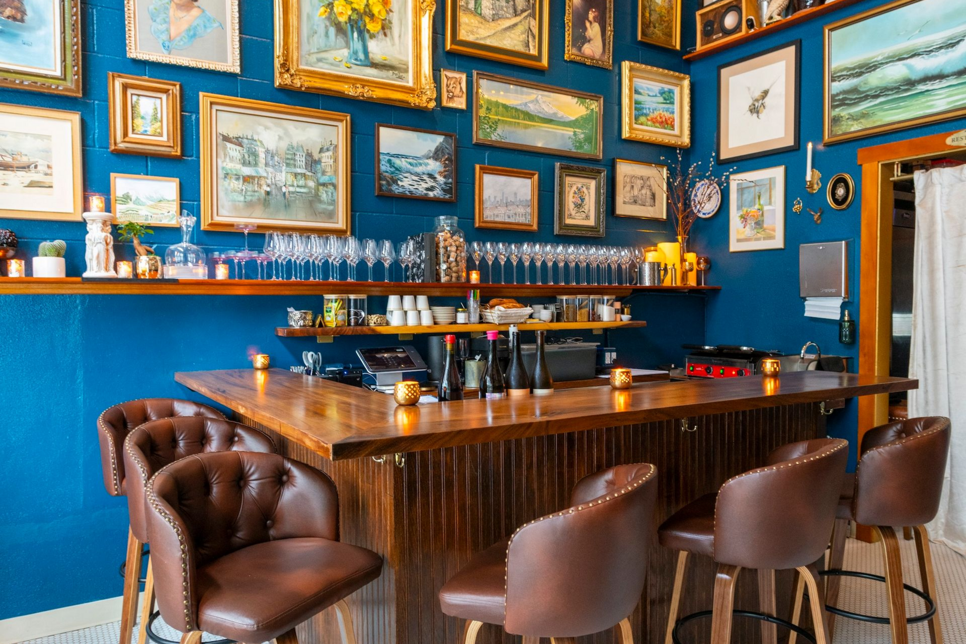 Cozy bar with blue walls, framed art, wooden tables, and leather-and-wood bar stools under a vaulted ceiling