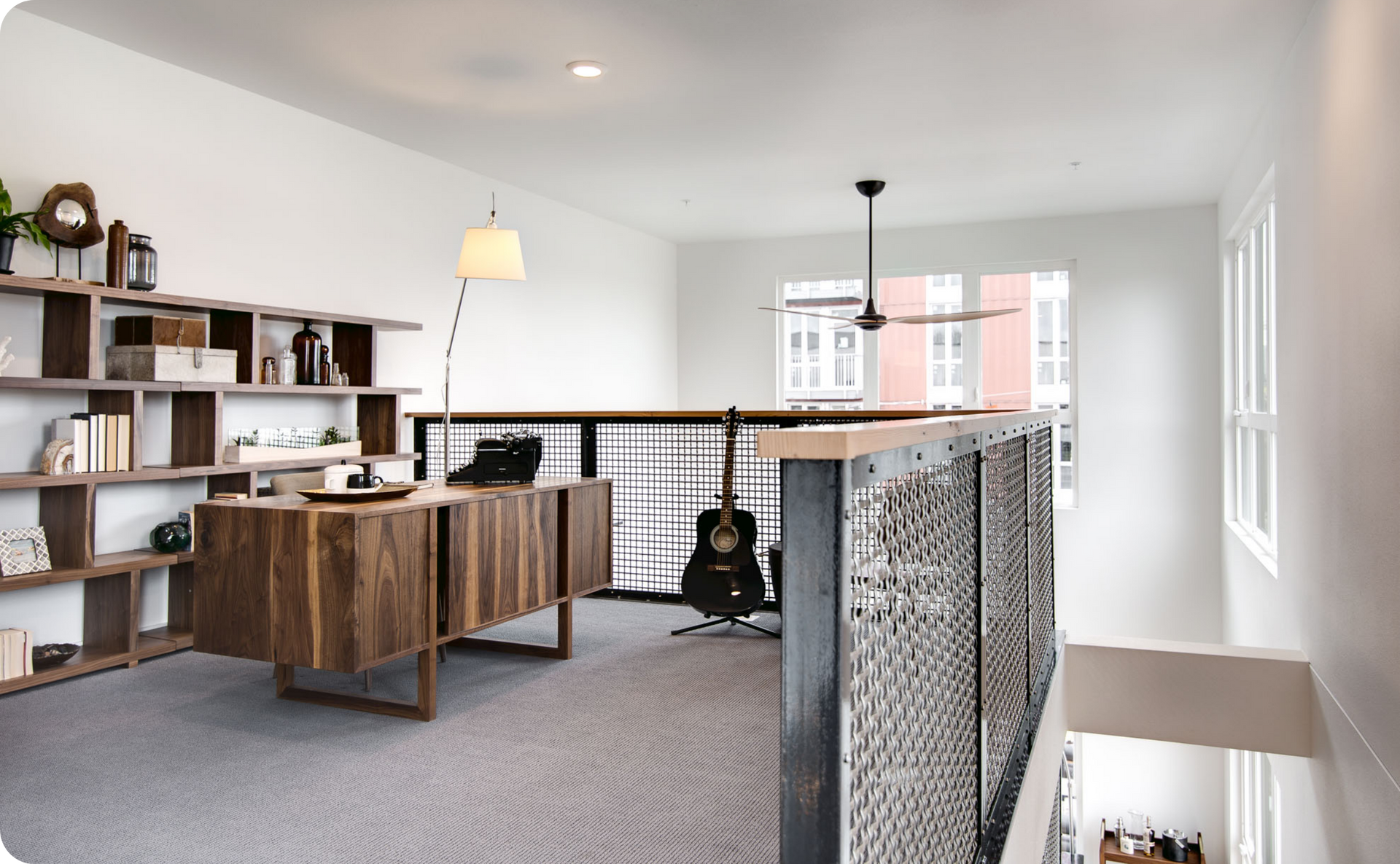 Modern home office with wooden desk, bookshelves, patterned rug, and a guitar by the railing.
