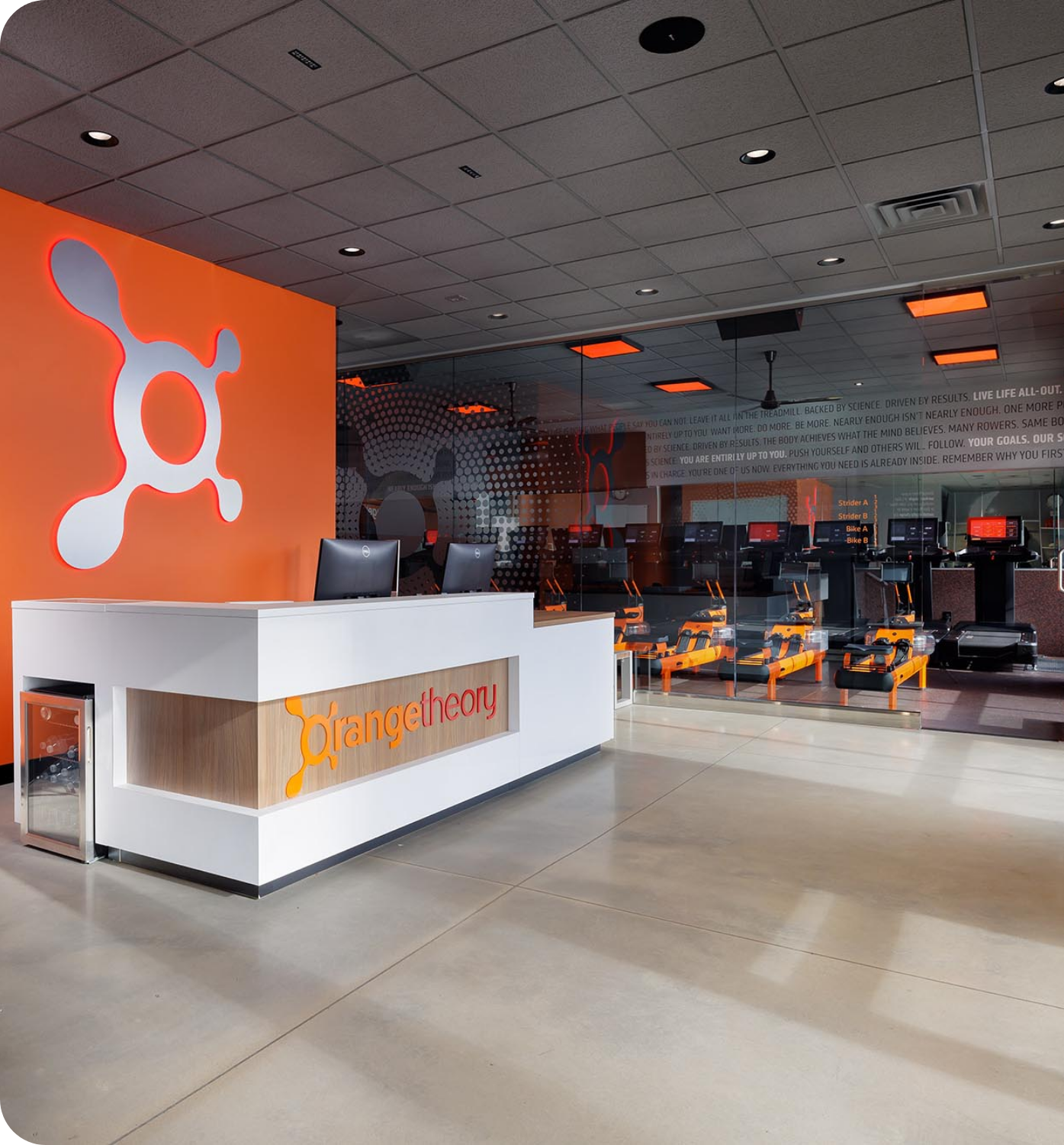 Modern office reception desk at Orangetheory Fitness with orange logo, glass-walled workspace, and polished concrete floor