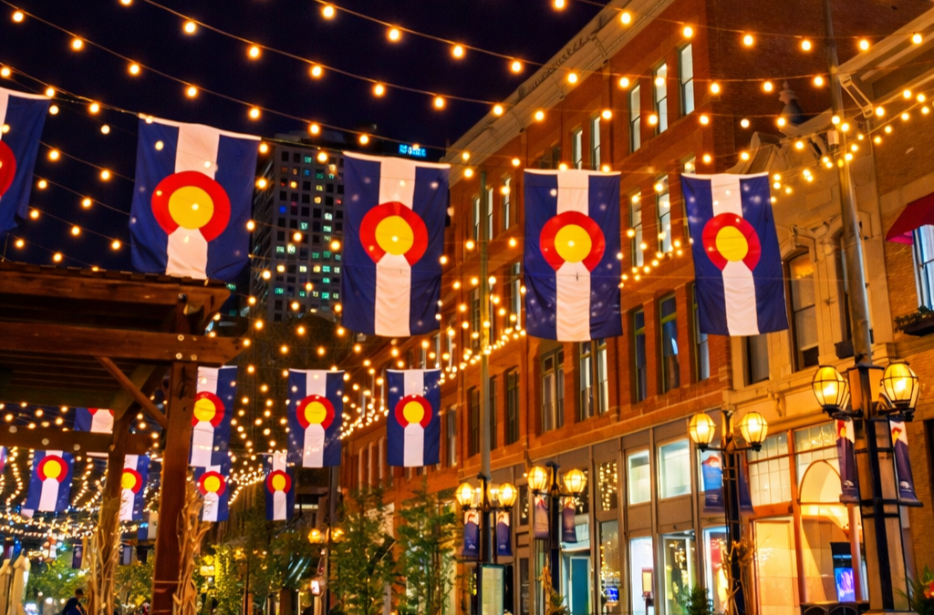 Colorado state flags hang vertically above a Denver, CO street, illuminated by festive string lights at night.