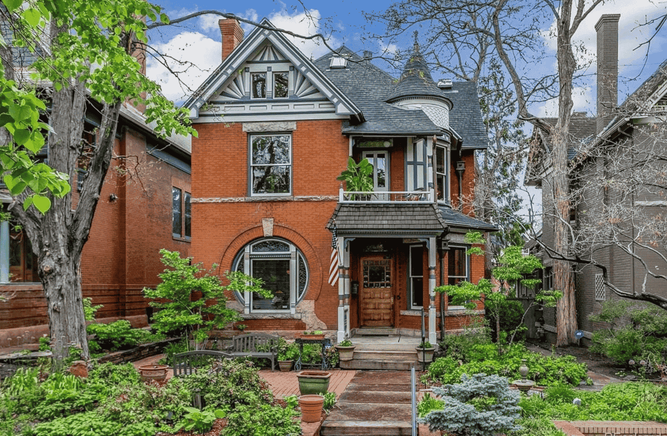 A large brick house with a porch and stairs is surrounded by trees and bushes.