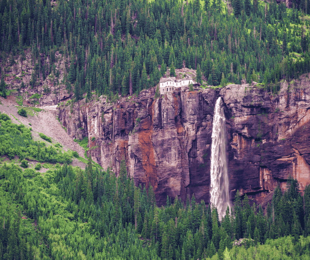 There is a waterfall on the side of a mountain surrounded by trees with a house on top