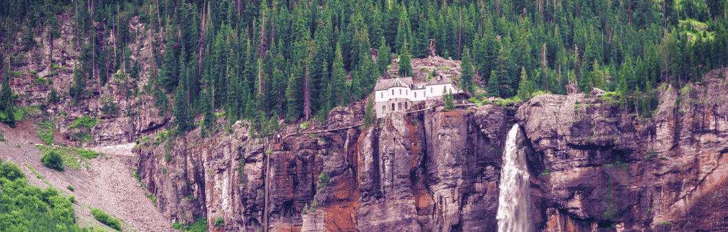 There is a waterfall on the side of a mountain surrounded by trees with a house on top