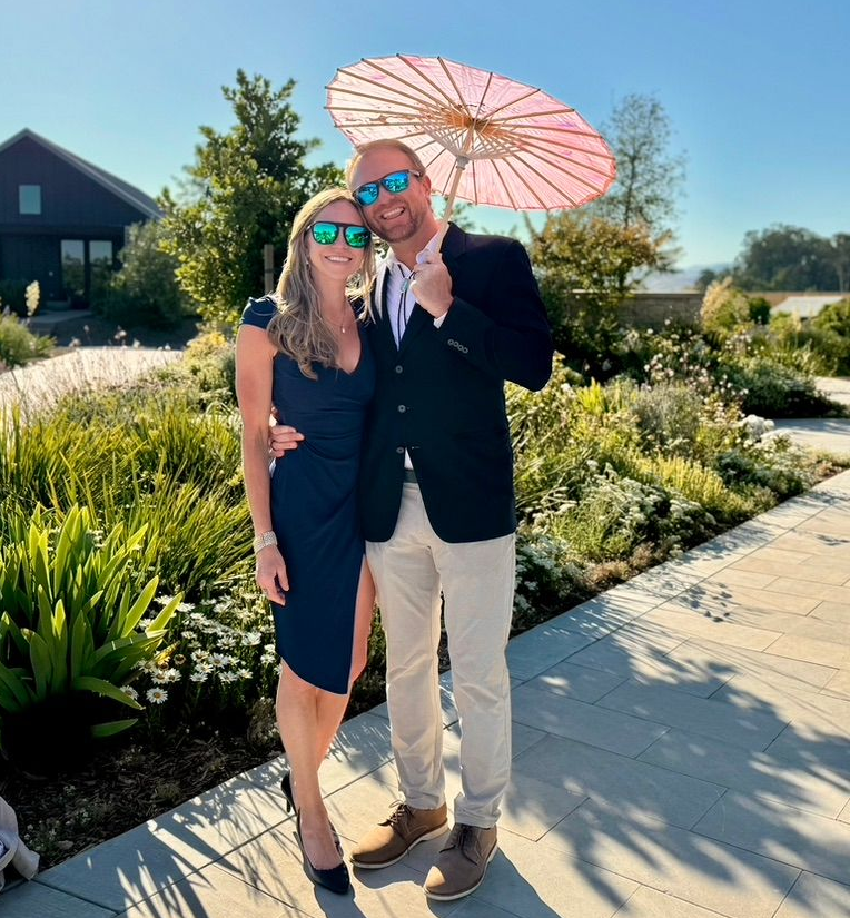 Will Ranking and wife smiling, posing outside with a pink umbrella, near greenery and a building.