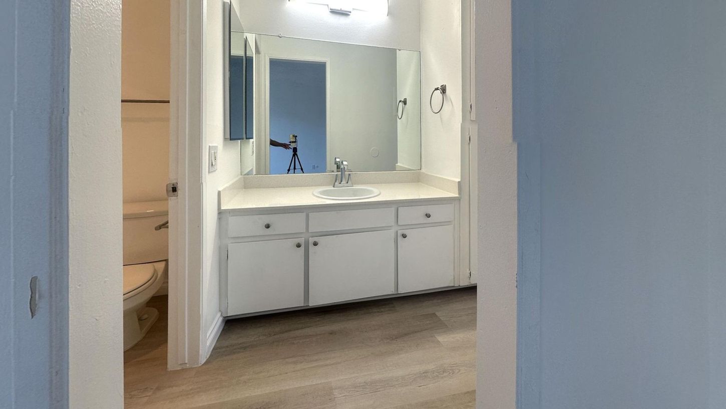 Bathroom with white vanity, sink, mirror, and toilet. Light blue walls and beige carpet.