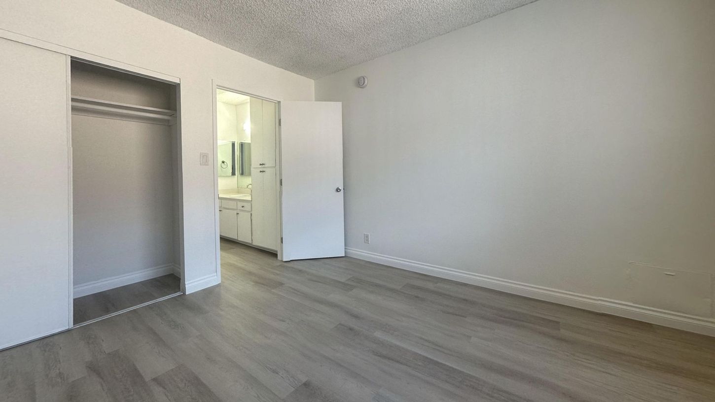 Empty bedroom with sliding closet doors, bathroom visible through an open door, and light wood flooring.
