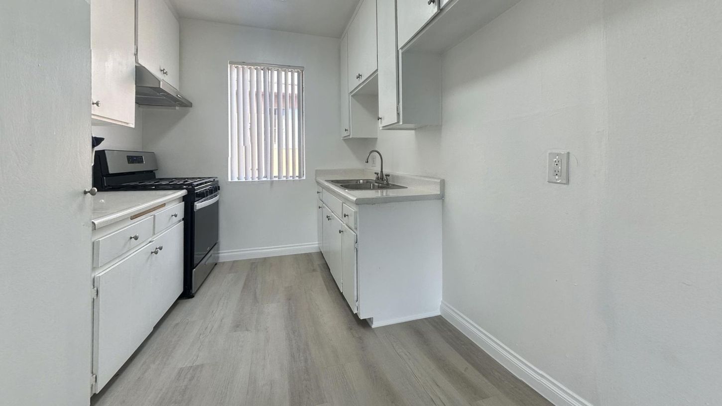 White kitchen with cabinets, sink, and stove. Light wood-look flooring and a window with blinds.