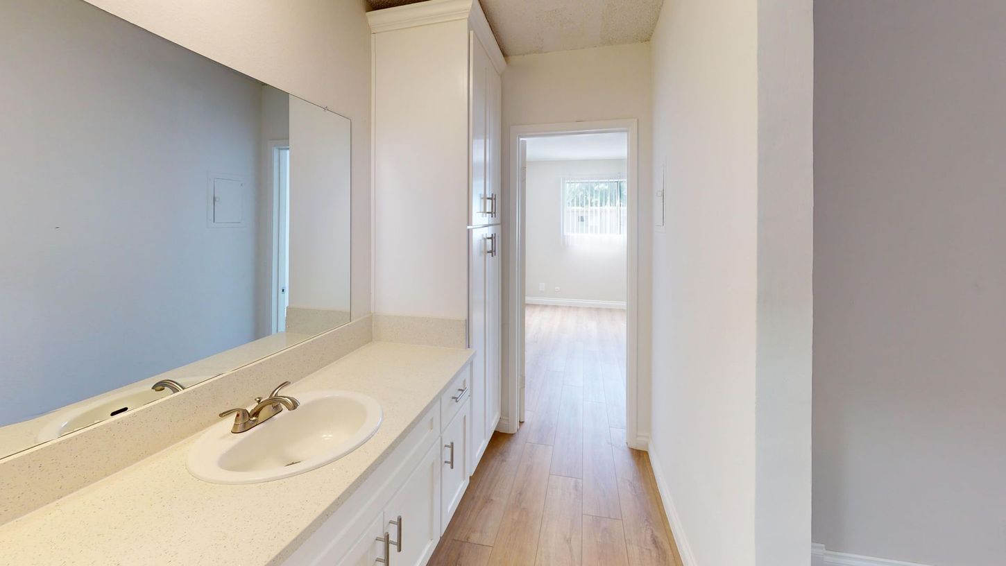 Bathroom with white cabinets, sink, mirror, and a hallway leading to a room with a window.
