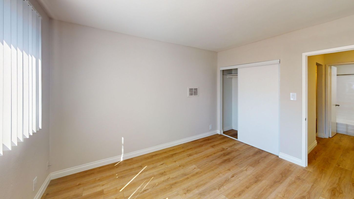 Empty room with wood-look floor, sliding closet door, window with blinds, and doorway to a bathroom.