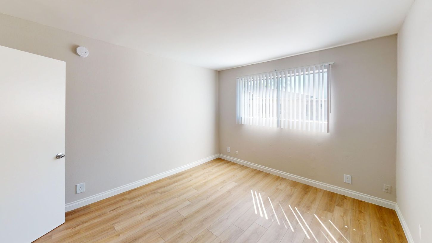 Empty room with a window, white walls, wood-look floor, and a closed white door.