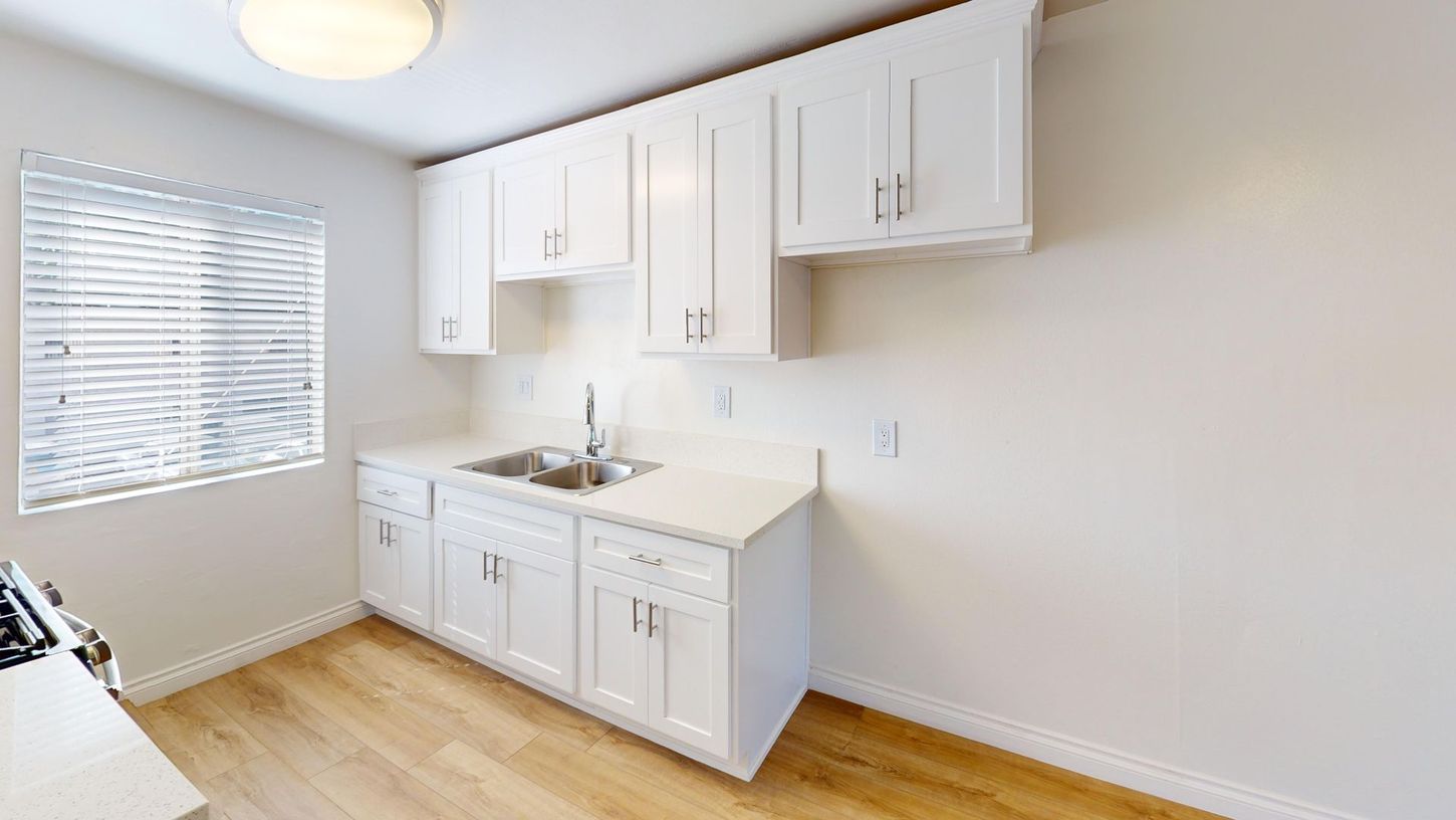 White kitchen cabinets, sink, window with blinds, and light wood flooring in a small room.