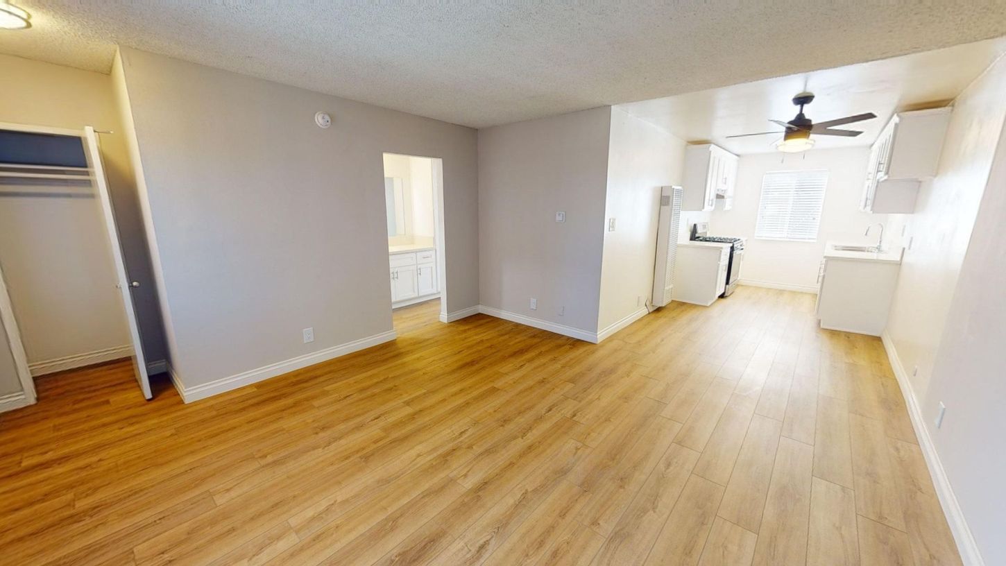 Interior of an apartment: Living area with light brown flooring, white walls, and a small kitchen.