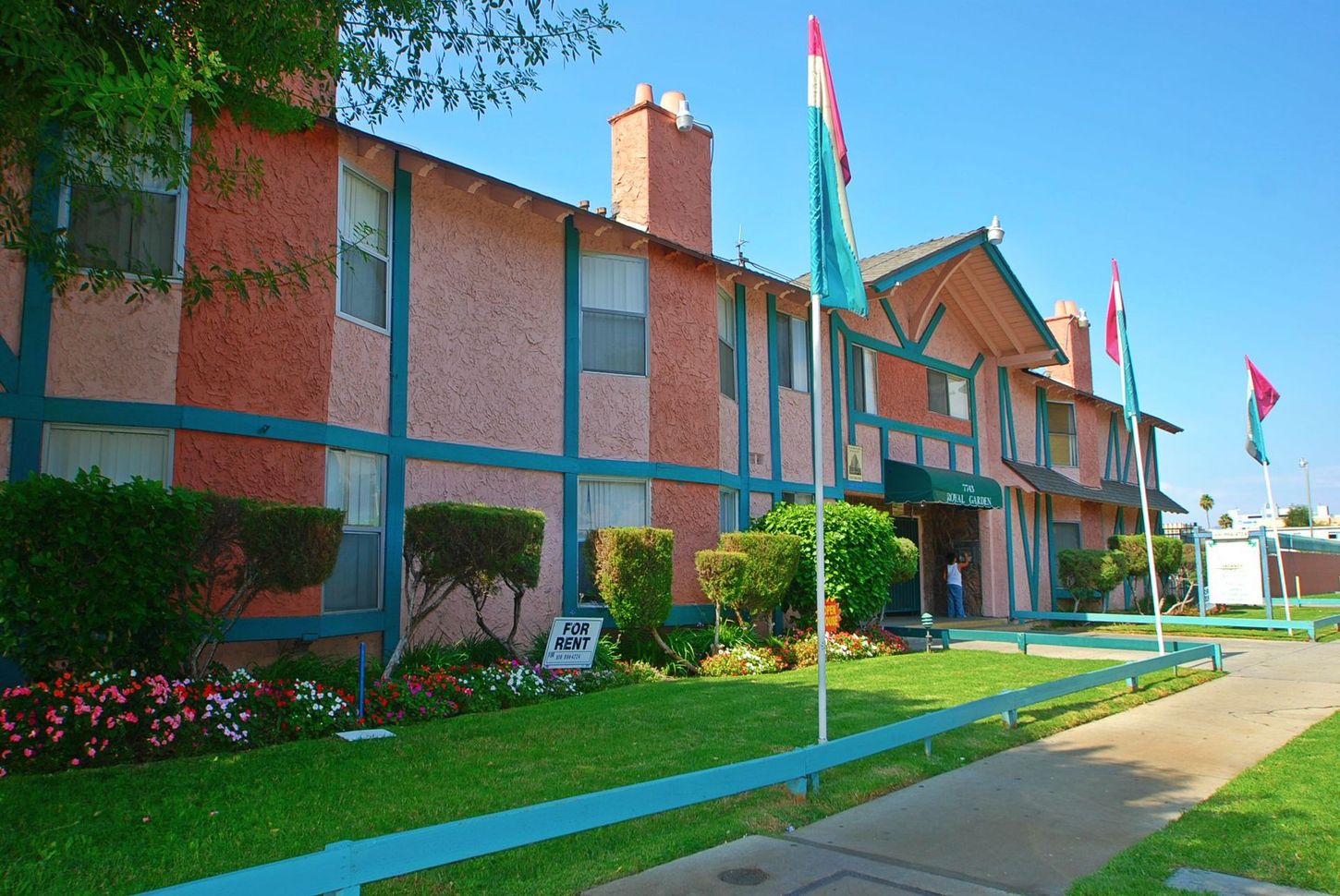 Two-story apartment building with pink exterior, teal trim, flags. Lush green lawn, blue sky.
