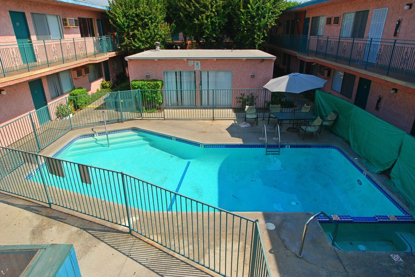 Pool area with light pink apartment buildings. A pool is in the center, with patio furniture and an umbrella.