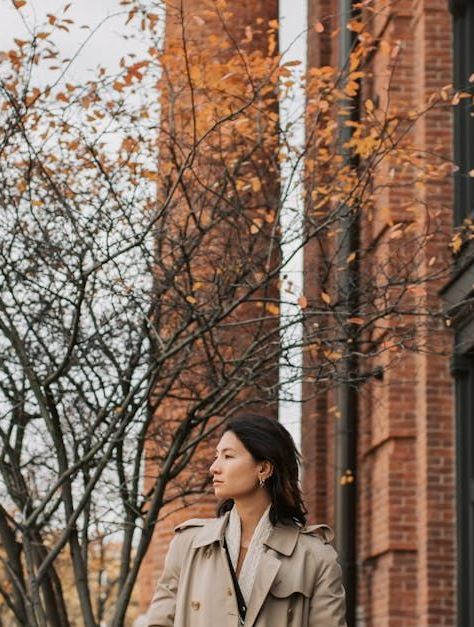 A woman in a trench coat is standing in front of a brick building.