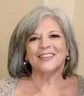 A woman is sitting at a table writing in a notebook next to a laptop.