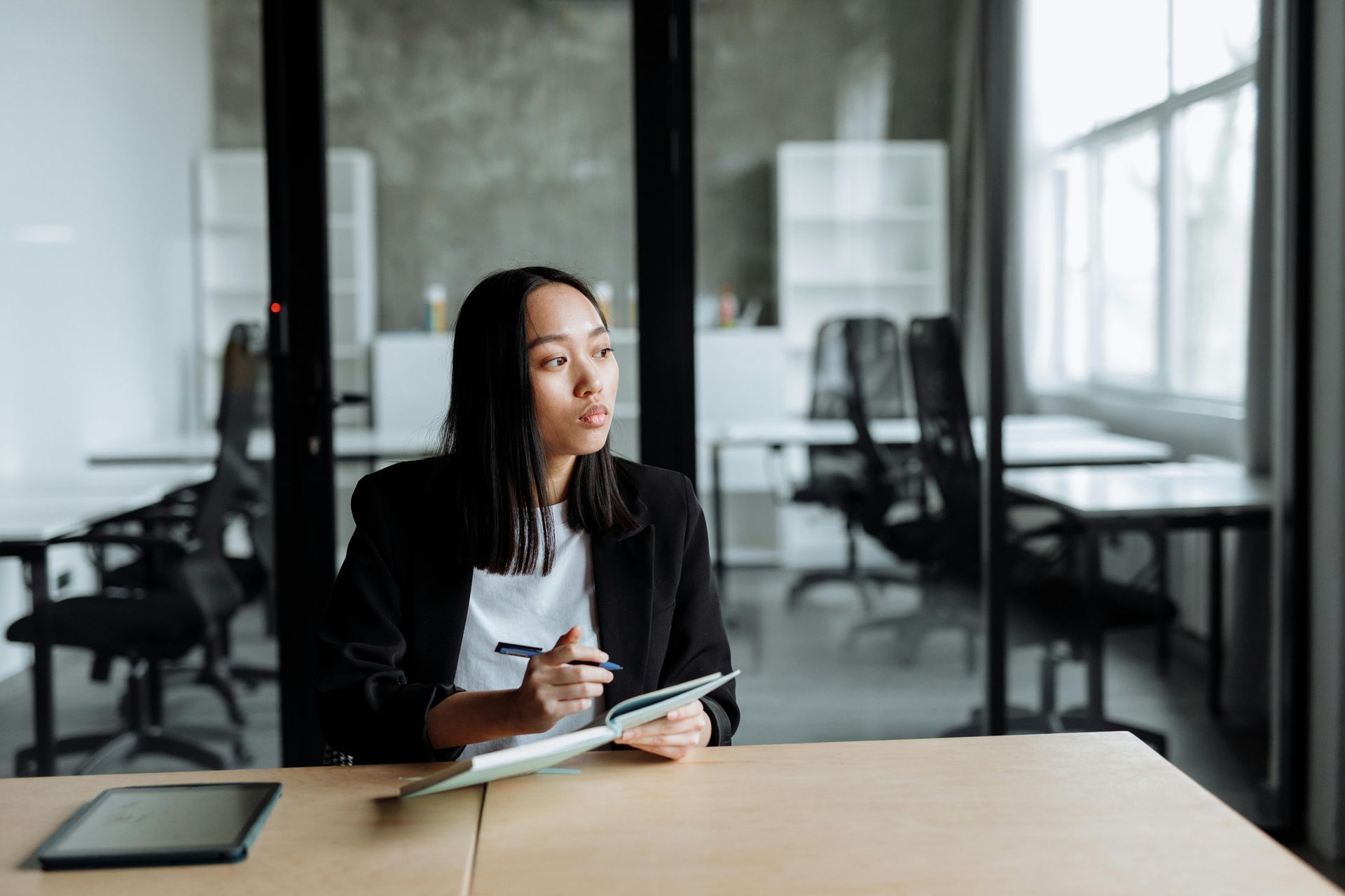 A woman is sitting at a table holding a pen and notebook and looking out the window.