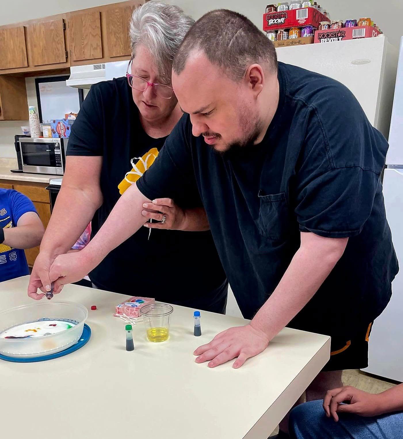 Woman helps a man with short dark hair at a table with colorful items, possibly crafting. They appear to be indoors, in a communal area.