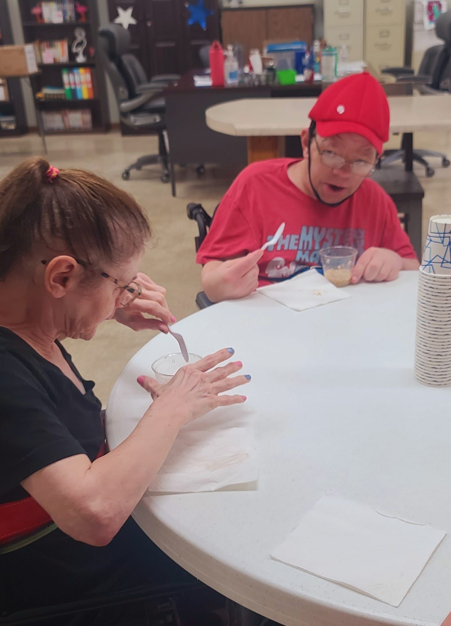 Two people at a round table, one cutting paper, the other eating. Both have disabilities and are indoors at a facility.