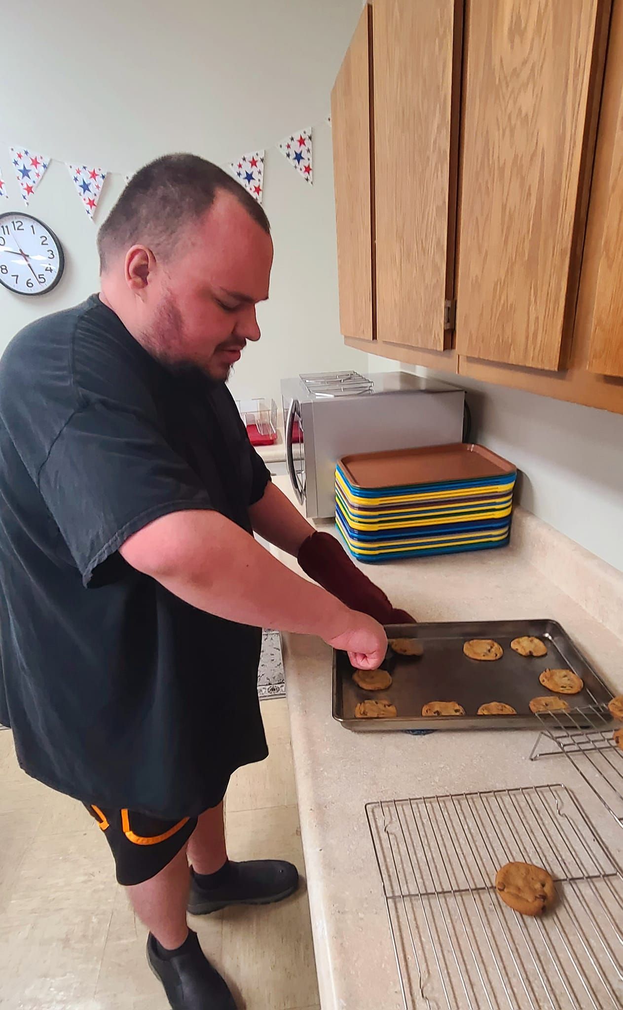 Man in black shirt and shorts removes cookies from a baking sheet with a spatula. He's in a kitchen with cabinets, oven mitts, and a cooling rack.